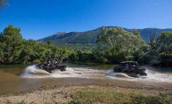 Two off-road buggies driving fast through a river with water splashing, mountains and blue sky in the background
